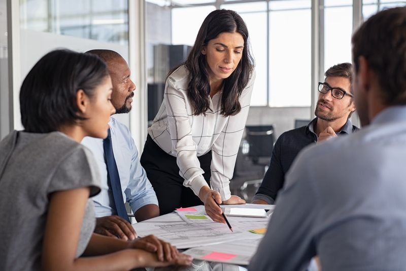 Photo of coworkers seated around a conference table. A woman is standing.